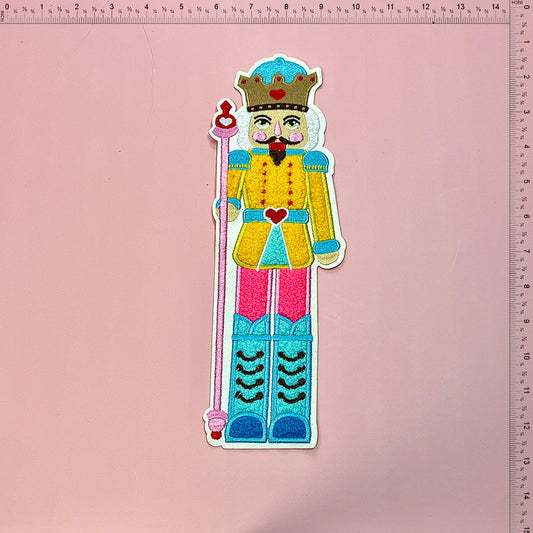 A vibrant, embroidered nutcracker figure dressed in bright colors, complete with a crown and staff, perfect for festive decor.Patches