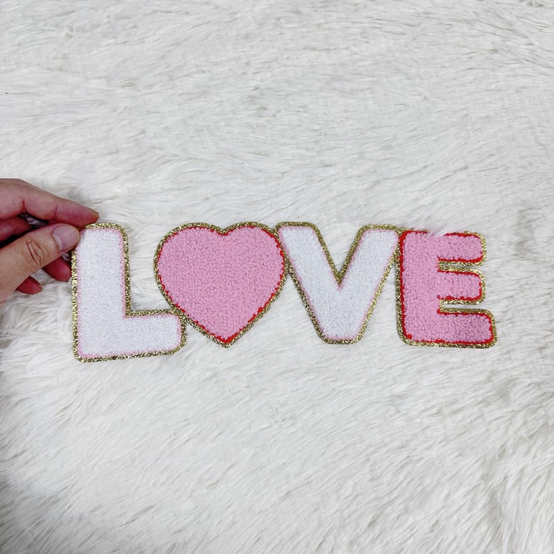 A charming decorative piece spelling "LOVE" with a pink heart, featuring soft textures and golden trim for a cozy touch.Patches